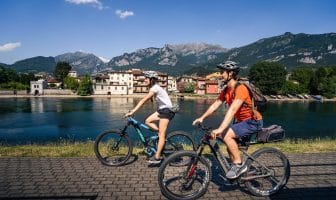 Persone in biciletta lungo la pista ciclabile l'Anello dei Laghi con sullo sfondo Pescarenico e il Monte Resegone